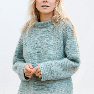 Une femme aux longs cheveux blonds porte un pull ample en tricot bleu clair et se tient debout sur un fond blanc uni. Son visage est partiellement visible et elle tient ses mains jointes devant elle.