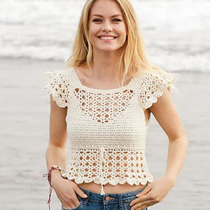 Une femme souriante se tient sur une plage, vêtue d'un haut à manches courtes couleur crème, crocheté, aux bords festonnés et aux motifs façon dentelle. Une inspiration parfaite si vous cherchez un modèle de haut crochet gratuit à télécharger. L'océan et les vagues se fondent en arrière-plan.