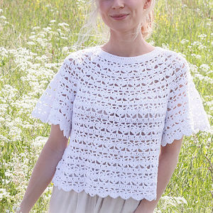 Femme debout dans un champ de fleurs sauvages, portant un haut à manches courtes en crochet blanc avec un ourlet et des manches festonnés, réalisé à partir d'un motif complexe et aéré - une inspiration parfaite si vous recherchez un modèle de haut crochet gratuit à télécharger.