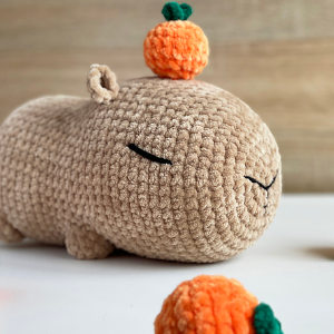 Une peluche de capybara crochetée, les yeux fermés, repose sur une surface. Un fruit orange crocheté avec une feuille verte repose sur sa tête, et un autre fruit similaire est au premier plan : une inspiration parfaite pour des modèles de crochet débutant gratuits.