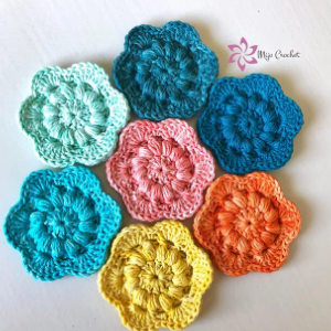Sept motifs floraux colorés au crochet sont disposés en cercle sur fond blanc. Présentés dans des modèles de crochet débutant gratuits, les fleurs sont bleues, bleu sarcelle, turquoise, aigue-marine, roses, orange et jaunes, avec le logo Myp Crochet en haut à droite.