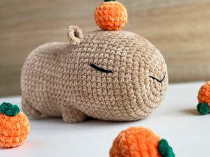 Un capybara en peluche crocheté, les yeux fermés, repose sur une surface blanche. Entouré de petits fruits, il porte un petit fruit orange en peluche sur sa tête. Parfait pour ceux qui recherchent des modèles de crochet débutant gratuits, l'arrière-plan est légèrement flou.