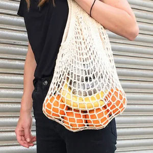 Une personne vêtue de noir tient un sac d'épicerie en filet réutilisable, probablement inspiré des modèles de crochet débutant gratuits, rempli de fruits, dont des oranges et une banane, devant un arrière-plan en métal.