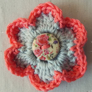 Une fleur crochetée inspirée des modèles de fleur au crochet faciles, avec des pétales bleu clair bordés de fil rose et un bouton à motif floral au centre, présenté sur un fond de tissu beige.