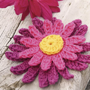 Une fleur crochetée avec des pétales roses et violets superposés et un centre jaune repose sur du bois blanc patiné, mettant en valeur l'un des modèles de fleur au crochet faciles, tandis qu'une vraie fleur magenta est partiellement visible en arrière-plan.