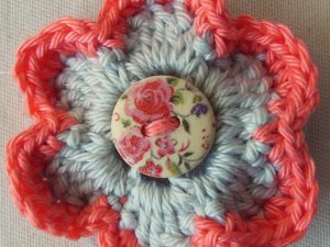 Une fleur crochetée aux pétales bleu clair bordés de fil rose, ornée d'un bouton rond orné d'un motif floral cousu au centre. Parfaite pour ceux qui recherchent des modèles de fleurs au crochet faciles. Le fond est un tissu uni clair.