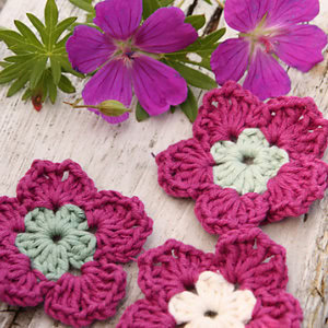 Trois modèles de fleurs au crochet faciles en fil rose et vert reposent sur une surface en bois blanc rustique, à côté de deux vraies fleurs violettes et de feuilles vertes.