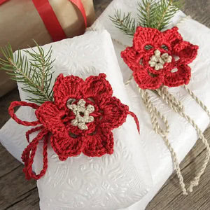 Deux cadeaux blancs emballés dans du papier gaufré sont décorés de modèles de fleurs au crochet faciles en rouge et beige, ainsi que de petites branches de pin, reposant sur une surface en bois rustique.