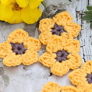Des fleurs crochetées jaunes et violettes, inspirées des modèles de fleurs au crochet faciles, sont disposées sur une surface en bois rustique à côté de fleurs jaunes fraîches et de feuilles vertes.