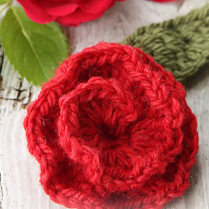 Gros plan d'une rose rouge crochetée avec une feuille verte, posée sur une surface en bois blanc rustique : une inspiration parfaite pour des modèles de fleurs au crochet faciles. Une vraie rose rouge et une feuille verte sont partiellement visibles en arrière-plan.