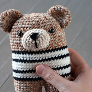 Une main tenant un adorable ours au crochet avec un corps beige et marron, des yeux en bouton noir, un museau crème et une chemise rayée noire et blanche, reposant sur une surface en bois grise.