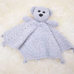 Une couverture de sécurité en forme d'ours en peluche douce et gris clair au crochet avec une tête et des bras d'ours attachés à une couverture triangulaire texturée, posée sur une surface duveteuse de couleur crème.