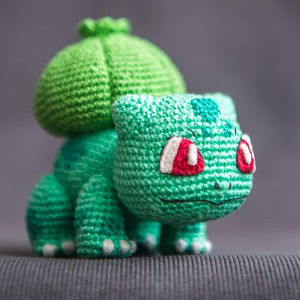 Un jouet en peluche Bulbasaur au crochet, avec du fil vert clair et foncé, des yeux rouges et blancs et une ampoule sur le dos, affiché sur un fond gris. Découvrez aussi des PDF gratuits d’amigurumis en français pour vos créations.