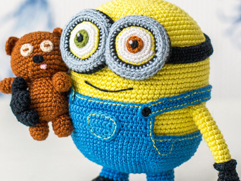 Un jouet minion jaune et bleu crocheté avec de grands yeux et un sourire cousu tient un petit ours en peluche marron crocheté, parfait pour les fans à la recherche de PDF gratuits d’amigurumis en français.