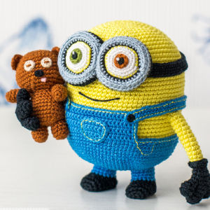Un Minion jaune au crochet en salopette bleue tient un petit ours en peluche marron, tous deux aux yeux expressifs, debout sur un fond clair. Découvrez ce modèle avec des PDF gratuits d’amigurumis en français pour les passionnés de crochet !.