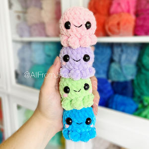 Une main tient une pile verticale de quatre pieuvres crochetées en rose, violet, vert et bleu, chacune avec des yeux noirs et un visage souriant. Des fils colorés sont visibles sur des étagères en arrière-plan — idéal pour trouver des PDF gratuits d’amigurumis en français.