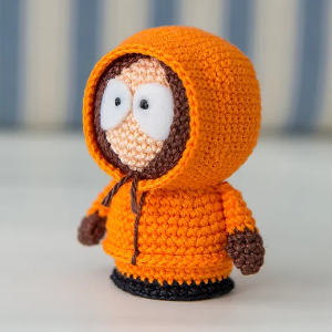 Une poupée au crochet aux yeux écarquillés, portant une veste à capuche orange avec une bordure marron, se tient sur une surface blanche avec un fond rayé flou.