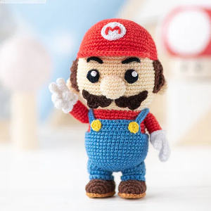 Une poupée au crochet ressemblant à Mario de la série de jeux vidéo Super Mario, portant un chapeau rouge avec un M, une chemise rouge, une salopette bleue et des gants blancs, se tient debout et fait signe d'une main.
