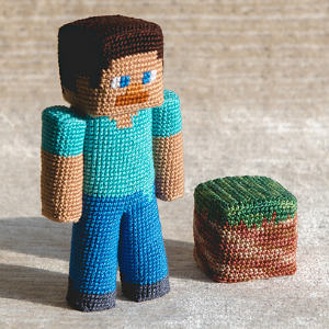Une figurine au crochet ressemblant à un personnage de Minecraft se tient à côté d'un bloc d'herbe et de terre au crochet sur un fond neutre.