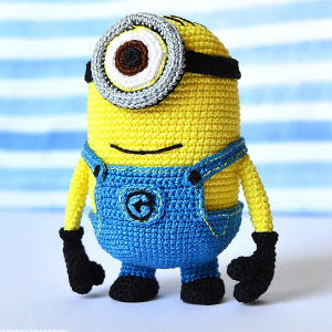 Une peluche au crochet ressemblant à un personnage de Minion, avec un grand œil, un corps jaune, une salopette bleue et des gants noirs, se tenant sur un fond rayé bleu et blanc flou.