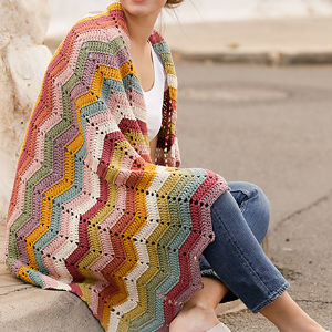 Une personne assise drape sur ses épaules une couverture colorée en crochet à carreaux et à chevrons. Vêtue d'une chemise blanche et d'un jean bleu, elle se détend dehors, sur un trottoir, près d'un muret de pierre.
