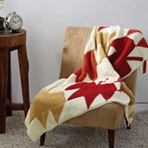 Une couverture en tricot confortable avec des motifs de feuilles géométriques rouges, dorés et crème, rappelant les motifs de crochet à carreaux, est drapée sur une chaise beige à côté d'une table d'appoint en bois avec un réveil sur le dessus.