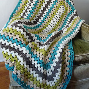 Une couverture carrée de grand-mère au crochet à carreaux colorés avec des rayures bleu sarcelle, vert, marron, beige et blanc drapée sur une chaise rembourrée verte.