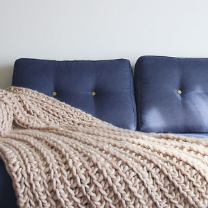 Une couverture épaisse en tricot de couleur crème et en crochet à carreaux drapée sur le siège d'un canapé bleu foncé avec des coussins capitonnés, posée contre un mur blanc uni.