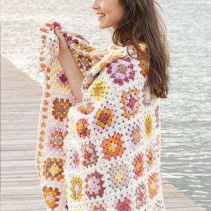 Une femme souriante se tient sur un quai en bois au bord de l'eau, enveloppée dans une couverture carrée en crochet à carreaux colorés avec des nuances de rose, d'orange, de jaune et de blanc.