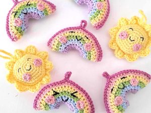 Porte-clés arc-en-ciel et soleil au crochet sur fond blanc. Les arcs-en-ciel ont des bordures roses et des détails floraux, tandis que les soleils sont jaunes avec des visages souriants et des joues roses.