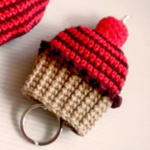 Un porte-clés au crochet en forme de cupcake, avec une base beige, un dessus rayé rouge et rouge foncé et un petit pompon rouge ressemblant à une cerise sur le dessus.