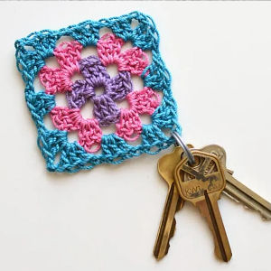 Un trousseau de clés est attaché à un porte-clés coloré en crochet, confectionné avec des fils bleus, roses et violets, selon un motif de carrés. Ce porte-clés carré et coloré repose sur une surface blanche.