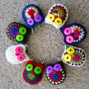 Sept porte-clés colorés en crochet en forme de hiboux aux yeux brillants sont disposés en cercle sur un tapis beige moucheté. Chaque hibou présente une combinaison unique de couleurs de fil et de détails charmants.