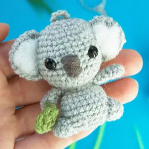Une main tient un petit porte-clés koala gris au crochet avec des yeux en perles noires, des oreilles blanches et une feuille verte attachée à son corps, sur un fond bleu.