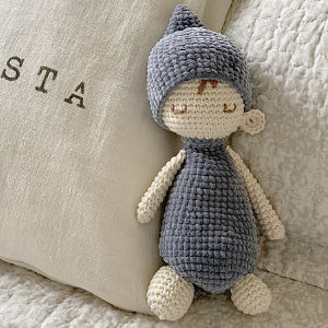 Une poupée au crochet avec un chapeau et un corps gris, des bras et des jambes de couleur crème et un petit visage brodé est posée contre des oreillers de couleur crème sur un couvre-lit texturé - l'une de nos charmantes poupées au crochet.