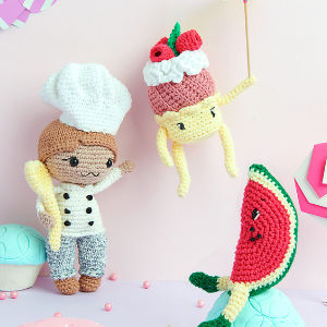 Trois poupées au crochet sont exposées : un chef tenant un rouleau à pâtisserie, un personnage de dessert jaune avec des baies et de la crème et une tranche de pastèque avec des bras et des jambes, le tout sur un fond rose pastel.