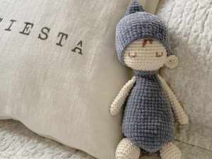 Une charmante poupée au crochet, vêtue d'un chapeau et d'une tenue gris, repose sur un lit à côté d'un oreiller de couleur crème brodé du mot SIESTA.