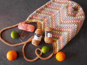 Un sac au crochet coloré avec explication gratuite est affiché sur une surface sombre avec deux écheveaux de fil, un crochet, une orange, deux citrons verts et une pelote de fil corail placés à proximité.