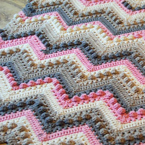 Gros plan d'une couverture bébé au crochet, ornée d'un motif chevron en zigzag, en fils crème, rose, marron et gris. La couverture présente des points texturés et des couleurs alternées.