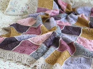 Une couverture bébé au crochet, composée de carrés de couleur rose pastel, or, violet et bleu avec une bordure en dentelle, est étalée sur un lit à motifs floraux.