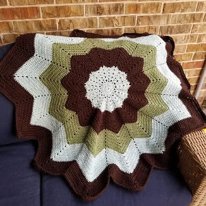 Une couverture bébé au crochet en forme d'étoile avec des anneaux alternés de fils blancs, marron et verts est drapée sur un canapé bleu foncé, avec un mur de briques et un panier en osier en arrière-plan.