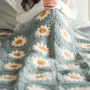 Une personne aux longs cheveux noirs est assise sur un lit, drapée dans une couverture bébé au crochet décorée de marguerites blanches et jaunes sur fond vert. Ses mains sont jointes et son visage est invisible.