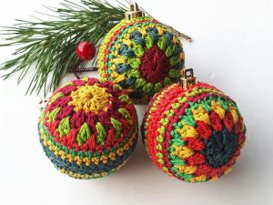 Trois décorations de Noël colorées au crochet en rouge, jaune, vert et bleu sont placées à côté d'une branche de pin avec une baie rouge sur fond blanc.