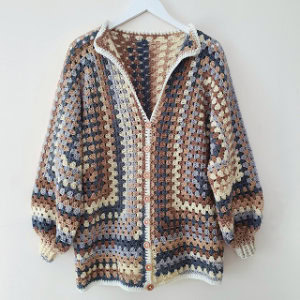 Un cardigan au crochet fait main inspiré des gilets au crochet pour femme, avec un motif patchwork de carrés de grand-mère en marron, beige, bleu et crème avec des manches longues et des boutons en bois, accroché sur un cintre blanc contre un mur uni.