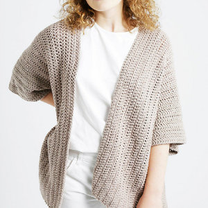 Une personne portant un gilet ample marron clair au crochet pour femme sur une chemise blanche et un pantalon blanc, debout sur un fond clair uni.