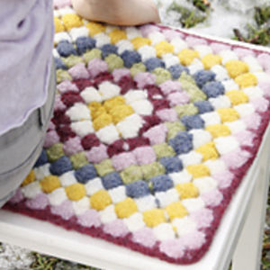 Un tapis au crochet coloré, fait à la main, avec un motif à carreaux en carrés violets, blancs, jaunes et bleus, réalisé à l'aide de modèles de crochet faciles et gratuits, est posé sur un banc blanc ; le genou et le bras d'une personne sont partiellement visibles.