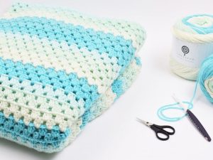 Une couverture au crochet pliée avec des rayures bleues, blanches et vert clair se trouve à côté d'une pelote de fil assorti, de petits ciseaux, d'un crochet et d'une aiguille à laine sur une surface blanche, parfaite pour les modèles de crochet faciles et gratuits.