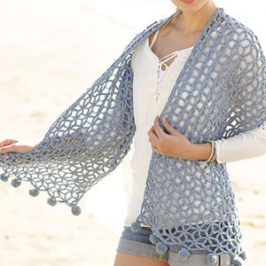 Une femme sur la plage porte un châle bleu clair au crochet ajouré, bordé de pompons, sur un haut blanc et un short en jean. Sa texture aérée et ajourée épouse les formes de la robe : une source d'inspiration idéale pour les modèles de crochet faciles et gratuits.