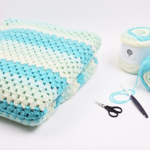 Une couverture pliée au crochet avec des rayures bleues, blanches et vert clair se trouve à côté d'une pelote de laine, d'un crochet et de petits ciseaux sur une surface blanche - une inspiration parfaite pour des modèles de crochet faciles et gratuits.