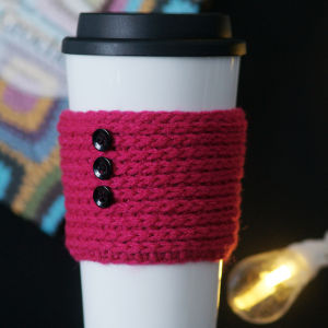 Une tasse à café de voyage blanche avec un couvercle noir est enveloppée dans une pochette rose texturée en tricot – parfaite pour les fans de modèles de crochet faciles et gratuits – fermée par trois boutons noirs. Un fond décoratif et une ampoule lumineuse sont partiellement visibles.